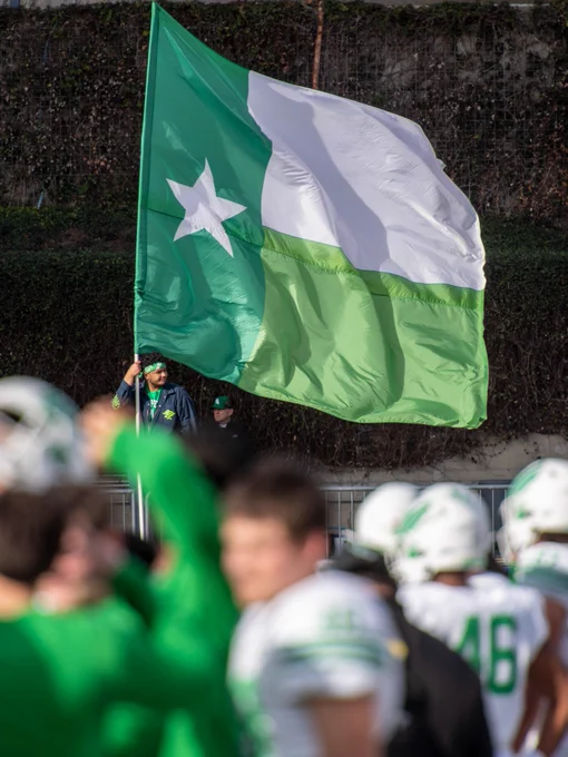 A photo of a person holding the UNT Battle Flag with Mean Green football players in the foreground.