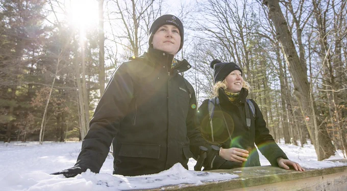 Two people in winter attire leaning on a wooden railing in a snowy park, smiling and looking into the distance, with sunlight streaming through trees behind them.