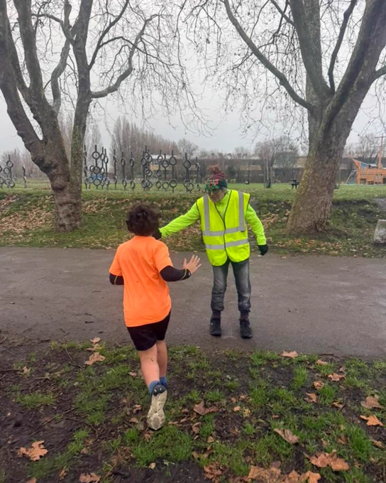 A junior parkrunner in an orange shirt running towards a volunteer for a high five.