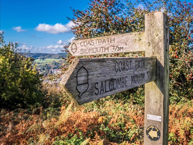 A wooden signpost on a sunny day points to "Sidmouth" and "Salcombe Mouth" along a coastal path. Lush greenery and blue sky in the background.