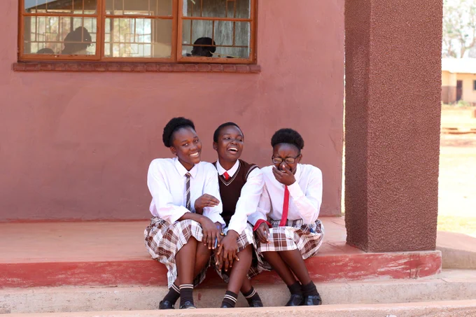 Three students in school uniforms sitting joyfully together on steps outside a building, laughing and sharing a moment.