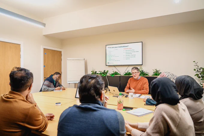 Group of people attentively listening to a presenter discussing a topic displayed on a screen in a modern meeting room.