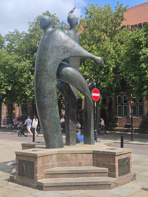 Bronze statue of 3 interlinked figures on a stone stepped plinth. Red brick building and trees in the background with passers-by. Summer day as blue sky and some cloud. People wearing summer clothes.