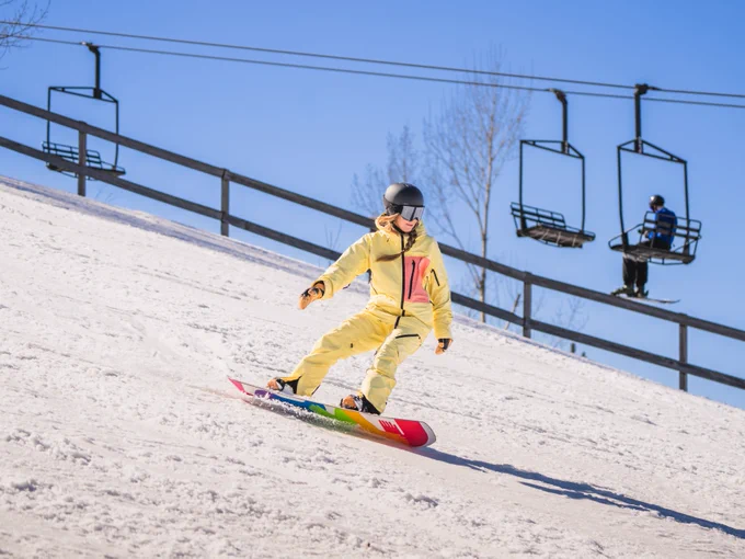 A person snowboarding down a slope with chairlifts in the background under a clear blue sky.