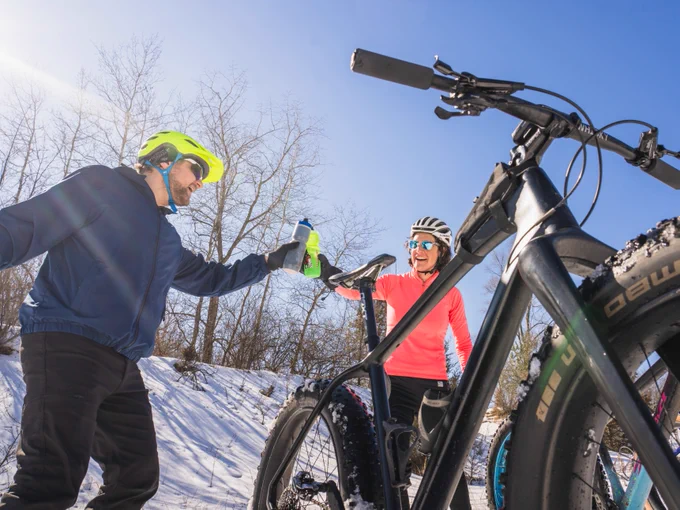 Two cyclists wearing helmets are taking a break and sharing a drink near their mountain bikes on a snowy trail under a clear blue sky.