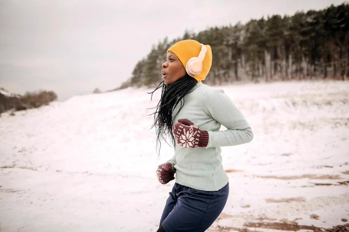 A person jogging on a snow-covered path, wearing winter attire including a yellow beanie, headphones, and mittens.