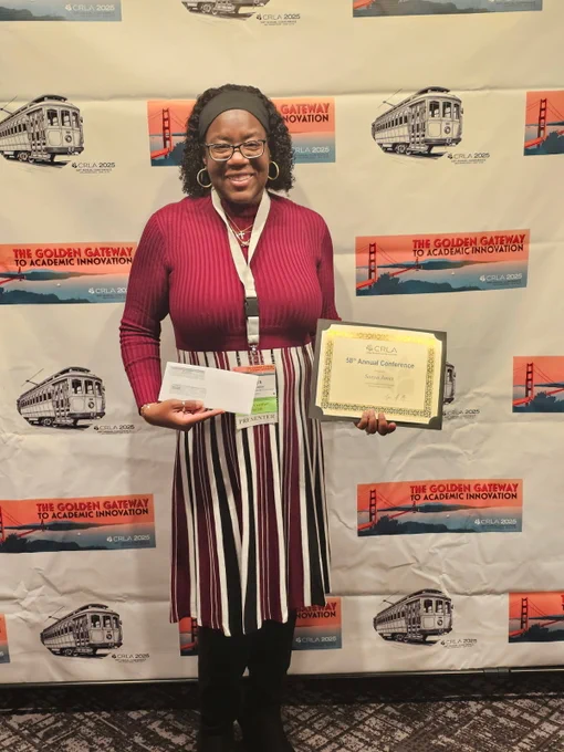 Person standing in front of a backdrop with logos, smiling and holding a certificate and an envelope.