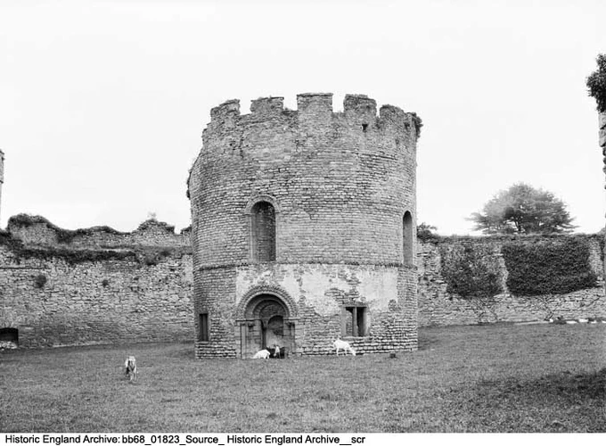 Black and white photograph showing a circular stone building set in a patch of grass bordered by a stone wall. The circular building has two principal stories and is topped with crenelation. A round headed doorway with zigzag mouldings gives access to the building. Goats or sheep graze on the grass.