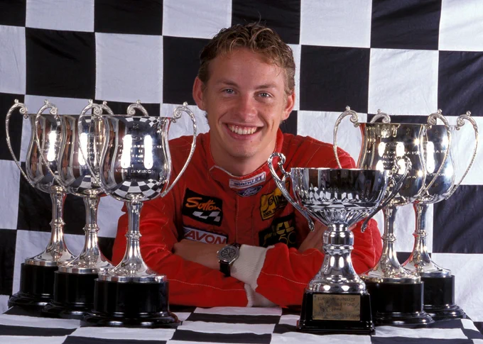 Jenson Button smiles in front of a chequered flag backdrop, arms folded on a table that also bears six handled cup style silver trophies. 1998.