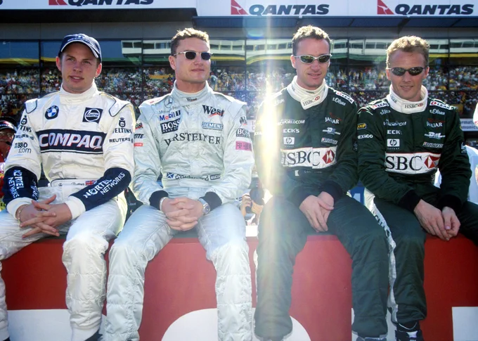 British drivers Jenson Button (white and dark blue Williams race suit), David Coulthard (off-white McLaren race suit), Eddie Irvine and Johnny Herbert (both dark green Jaguar race suits) sit in a row together on pit wall. All but Button wear fashionable sunglasses. 2000 Australian Grand Prix.