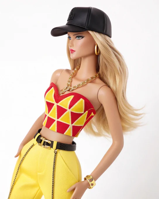 A Barbie doll with long blonde waves stands against a white background. She wears a red and yellow geometric print crop top with a square neckline and a black waist belt, paired with high-waisted yellow pants and a gold chain belt. Accessories include a black baseball cap with a gold logo, gold hoop earrings, and a gold bracelet.