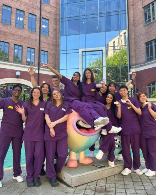 A group of final year dental students in purple scrubs holding up their DDU name badges and sitting on the colourful tooth statues outside Bristol Dental School.