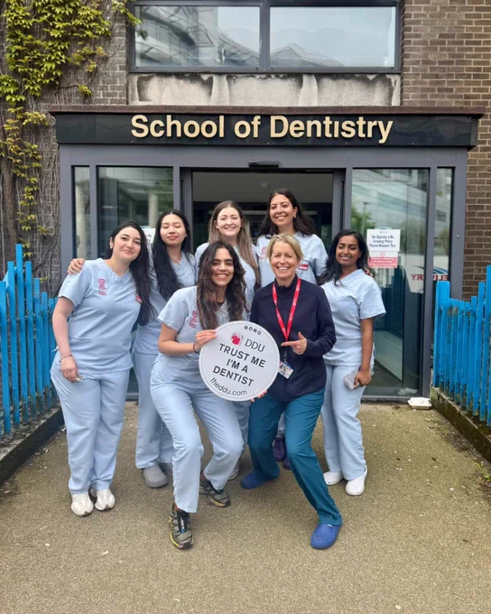 A photograph showing final year students and their tutor, holding an DDU branded 'Trust me, I'm a dentist' sign outside Belfast School of Dentistry.