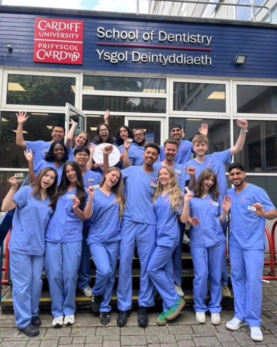 A group of final year dental students in light blue scrubs holding up their DDU name badges and celebrating outside of Cardiff School of Dentistry.