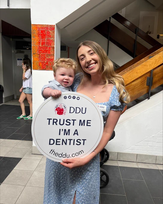 A final year dental student and her baby holding up the DDU-branded 'Trust me, I'm a dentist' sign inside a building with stairs in the background'.