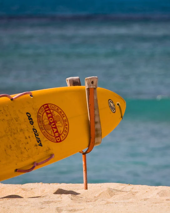 A yellow Ocean Safety lifeguard rescue board propped up in sand with a calm blue sea in the background.