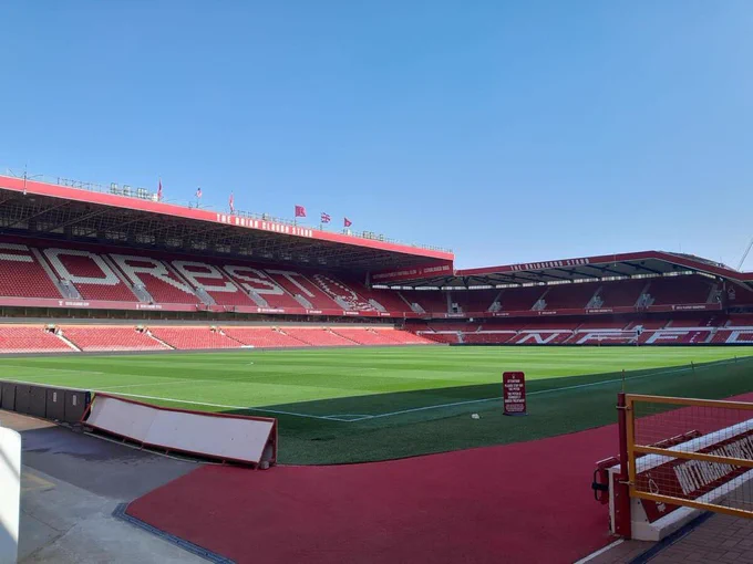 Photo of an empty City Ground stadium, the home of Nottingham Forest FC. The Brian Clough stand can be seen on the left of the shot whilst the adjoining Bridgford stand wraps around to the top right of the photo. The viewpoint looks directly to the corner where the two stands meet and the green pitch is illuminated with sunlight.