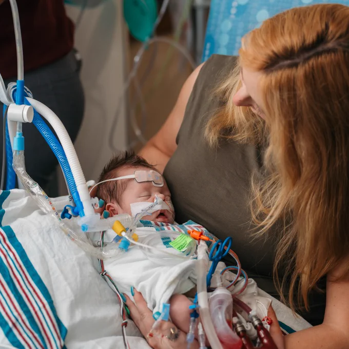 A holds holds a small infant connected to medical equipment in a hospital setting. 