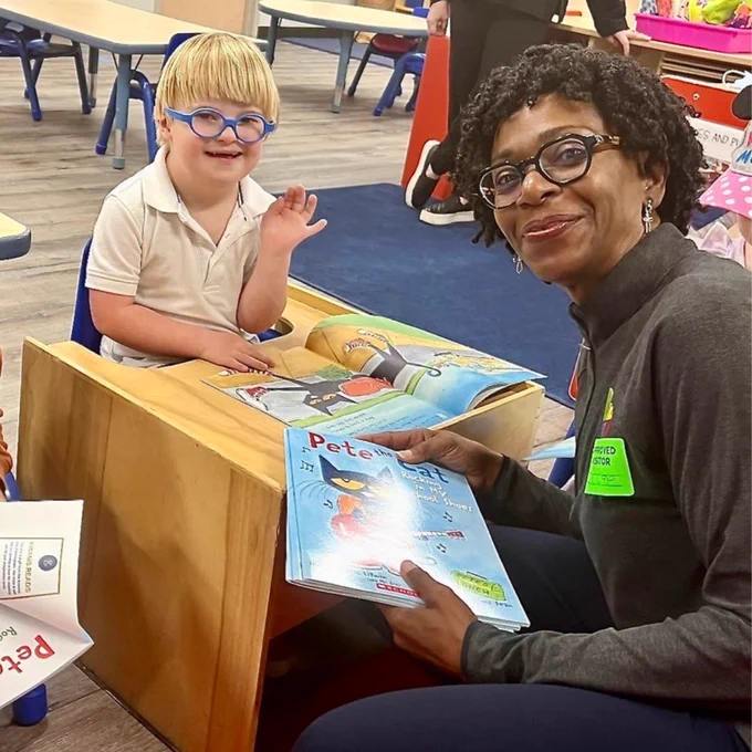 Child wearing blue glasses sits at a small table and waves while a teacher holds an open picture book beside him.