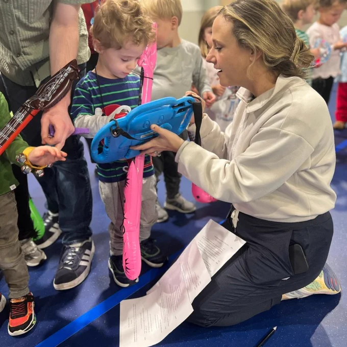 Teacher kneels on the floor and helps a young child use a AAC Device while other children stand nearby in a classroom.