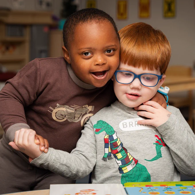 Two young children sit side by side at a table, holding hands and looking toward the camera.