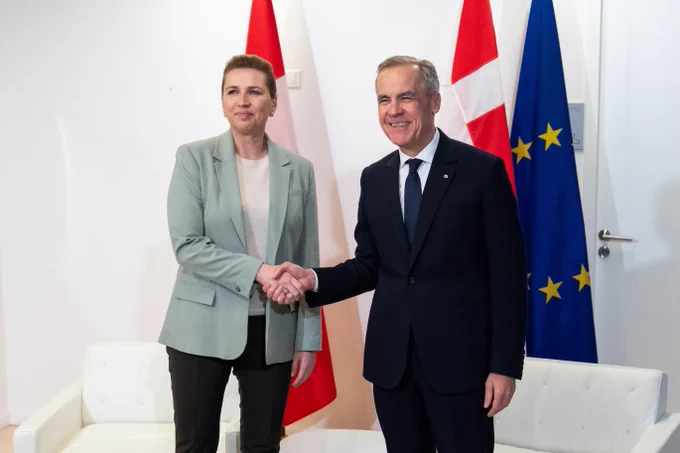 Prime Minister Mark Carney shakes hands with Denmark’s Prime Minister Mette Frederiksen during a bilateral meeting in Paris. The flags of Canada, Denmark, and the European Union are displayed behind them. 