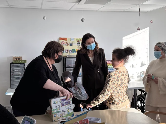 Photographie de la ministre aux côtés d’enfants et d’adultes. Nous apercevons des boites en plastiques et des dessins à l’arrière plan.