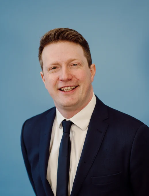Man wearing a dark navy suit with a white shirt and black tie against a plain blue background.