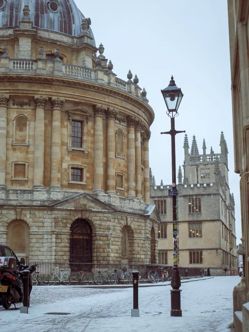 Snow gently falls on Radcliffe Camera and surrounding historical buildings in Oxford, complementing the serene winter scene with bicycles parked alongside the street and a street lamp in the foreground.