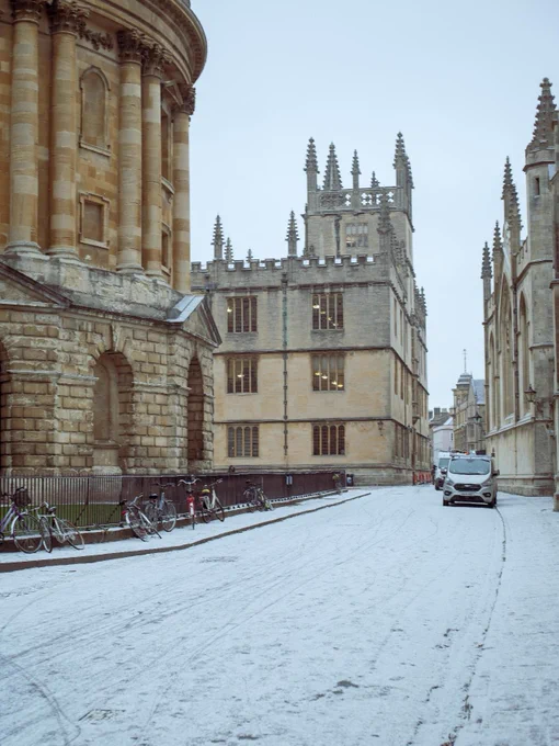 Snow-covered street in Oxford with the Radcliffe Camera to the left and historical buildings on the right, bicycles parked alongside.