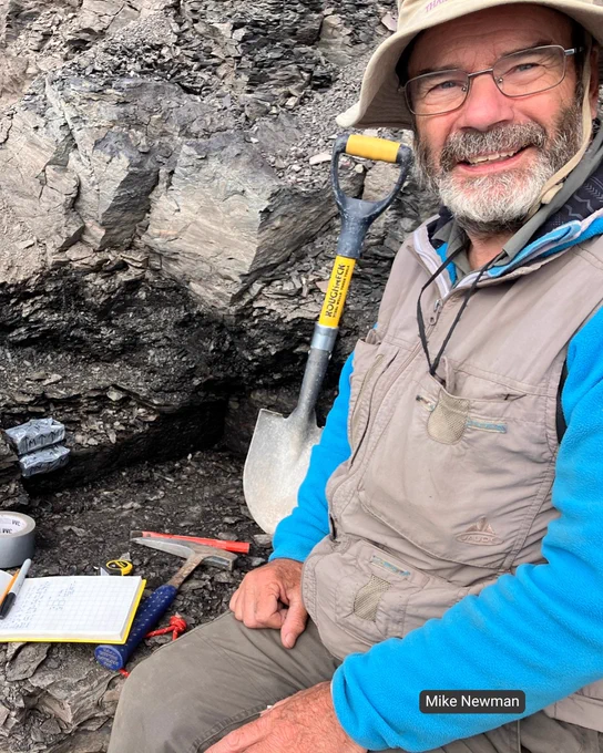 Professor John Marshal with glasses and a hat smiles at the camera while sitting beside a rocky outcrop. He is wearing a beige vest and blue sleeves. In front of him are geological tools including a hammer, measuring tape, and a notepad with notes. 