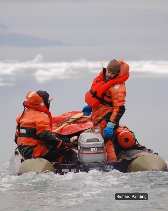 Two people in bright orange survival suits and life jackets are on a small inflatable boat. They are navigating icy waters, with icebergs visible in the background and a cloudy sky overhead. One person is handling the motor, while the other sits facing forward.