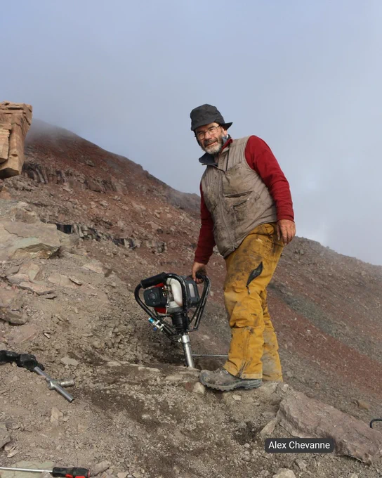 Professor John Marshall, in outdoor attire, operates a power drill on a rocky, barren mountainside. He wears a dark hat, glasses, and has a gray beard. The terrain is steep with loose dirt and stones. 