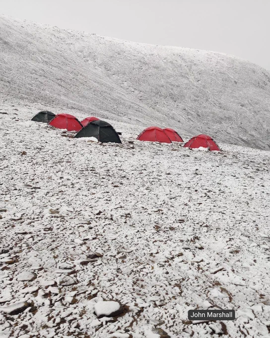 Red and black tents are pitched on a snowy, rocky slope under an overcast sky. The ground is thinly covered with snow, revealing many stones. The background shows a large, snow-dusted hill.