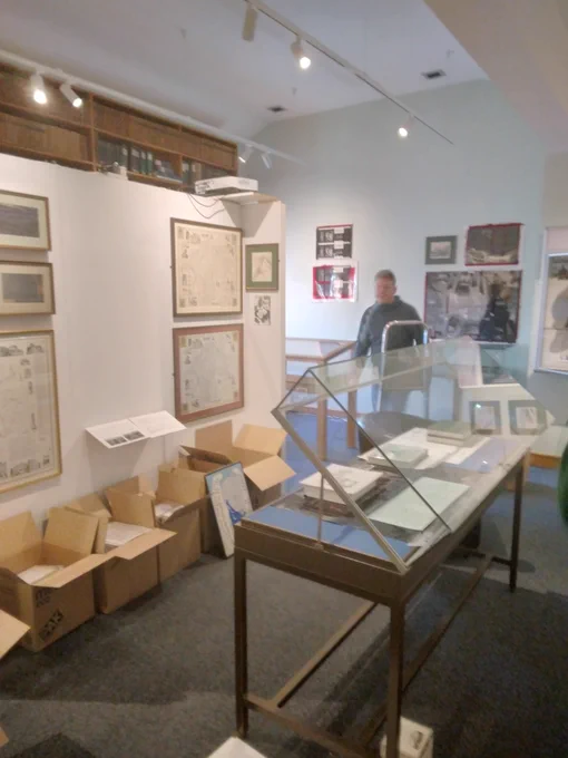An interior view of The Armitt's Alfred Wainwright exhibition featuring framed artworks and display cases. Boxes are seen in the background, indicating deinstallation.
