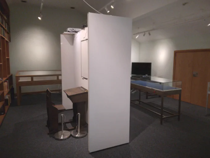 A view of an empty exhibition space featuring an old school desk and display cabinets.