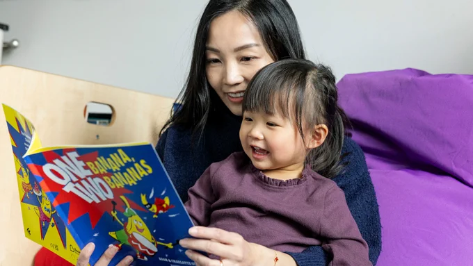 A mum and toddler sit on a purple bean bag reading a funny picture book together. They are cuddling and smiling. 