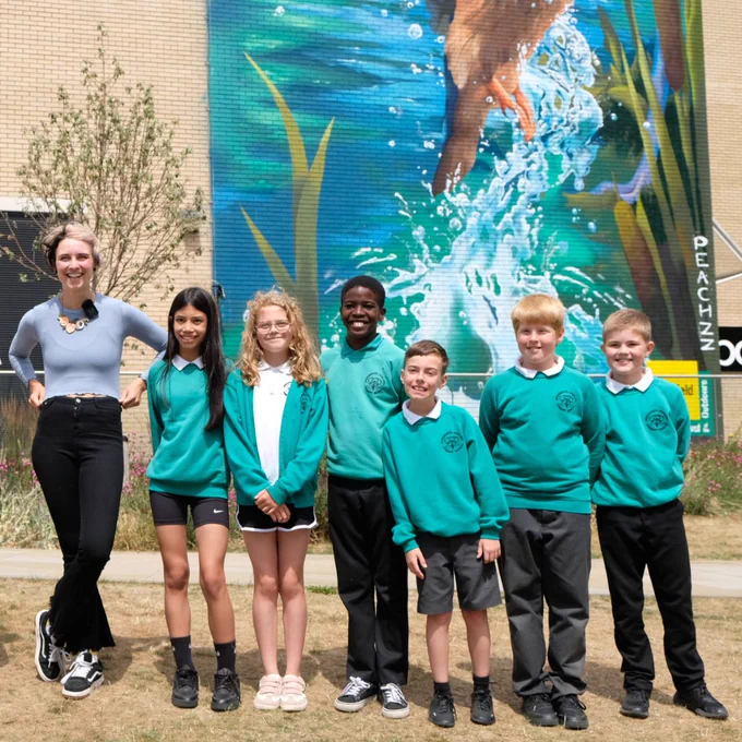 A group of six children in teal school uniforms stand outdoors with artist Peachzz. Behind them is colorful mural of a bird causing a splash in a canal.