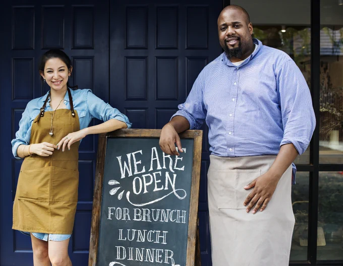 Two restaurant staff in aprons stand beside a chalkboard sign announcing brunch, lunch, and dinner service hours.