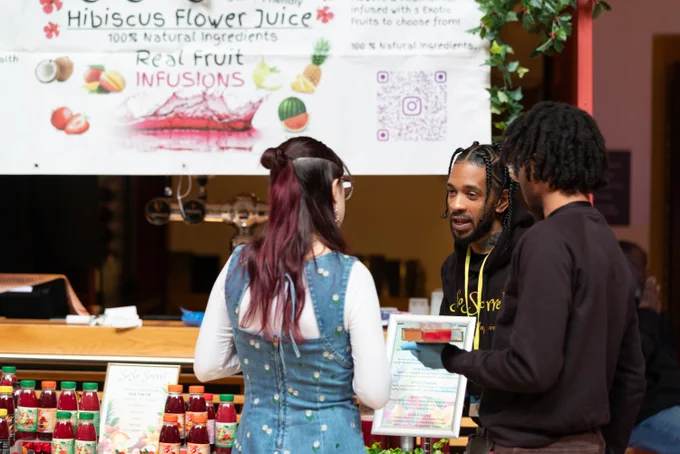 Three people stand at a market stall displaying colorful bottles of hibiscus flower juice and fruit infusions under a promotional banner.