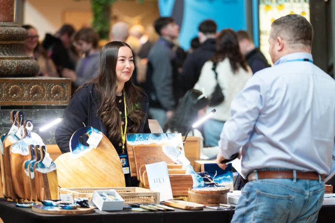Vendor showcasing wooden cutting boards and kitchenware at a busy market stall while a customer browses the items.