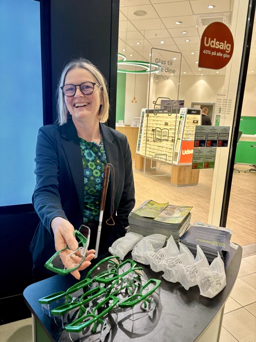 Diana står bag et højt bord og rækker et par beskyttelsesbriller frem. På bordet ligger beskyttelsesbriller og en pjece om synet.