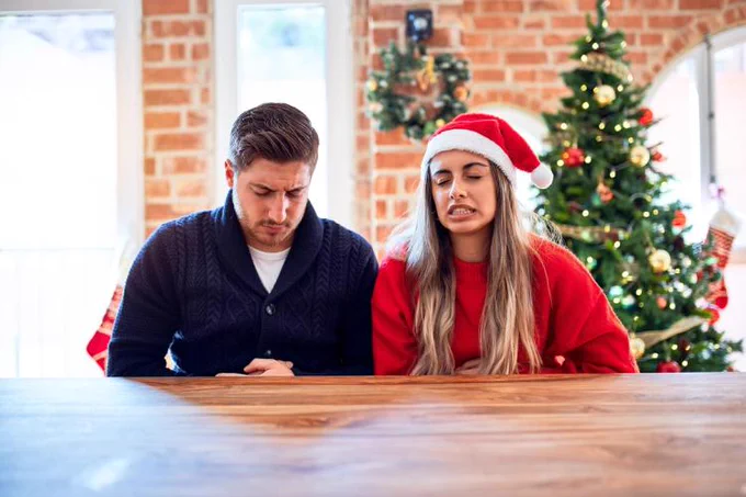 Two people sitting at a table, one wearing a Santa hat, with a decorated Christmas tree in the background.