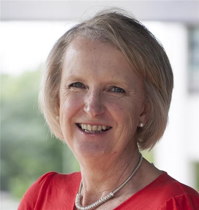 Professor Anna Horwood wearing a red top and silver necklace, standing outdoors with blurred greenery in the background.