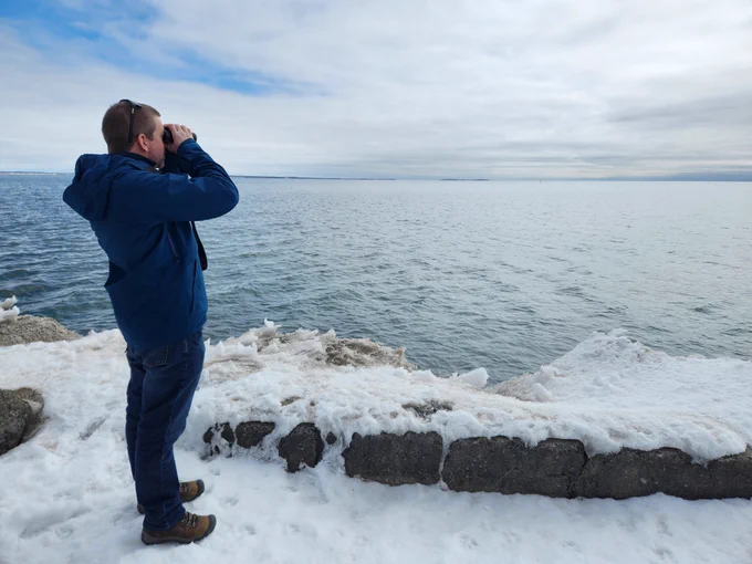 Person looking through binoculars standing beside a large lake with snow on the ground and a cloudy sky. 
