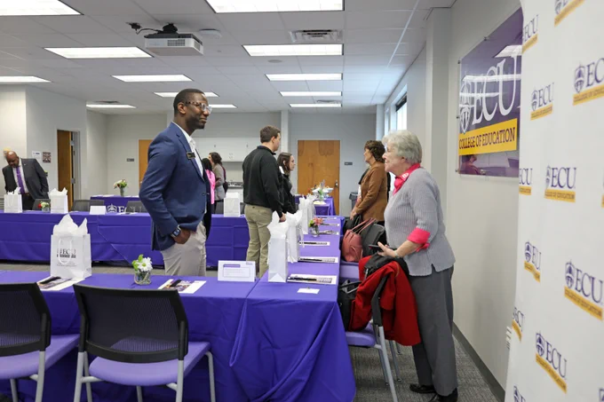 Individuals interacting at an East Carolina University event, featuring informational booths with purple tablecloths and ECU banners.