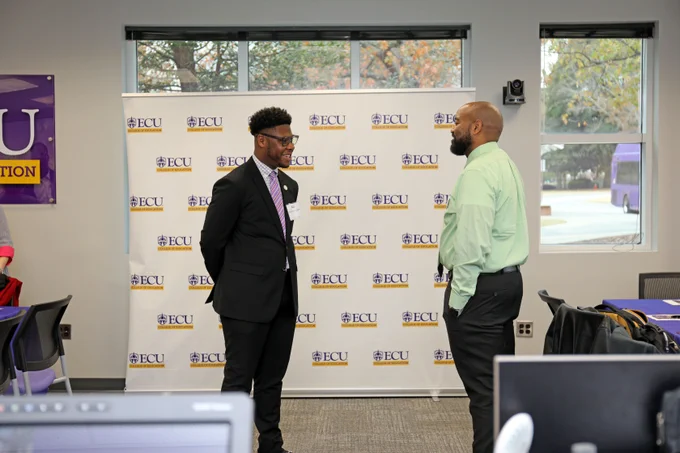 Two individuals conversing in front of a backdrop with the East Carolina University (ECU) logo.