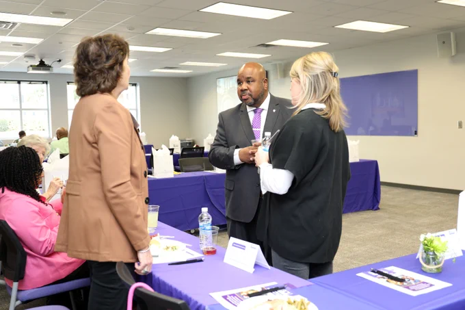 Three individuals are having a conversation in a meeting room with tables, chairs, and promotional materials visible around them.