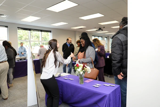Two people are shaking hands over a table at an indoor event, with others conversing in the background. The table is draped with a purple cloth and decorated with white flowers.
