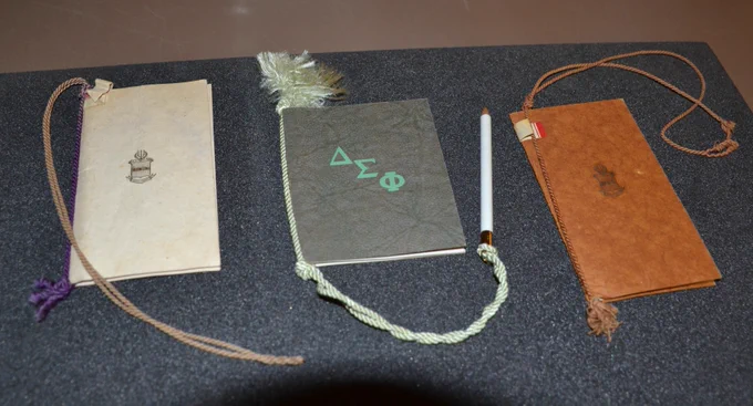 Three dance cards aligned on a foam pad. from left to right, a white one, a gray one and a brown one. all with twine attachment for a pencil. 